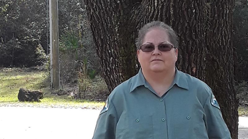 a woman with short hair and in a green uniform stand sin front of a tree